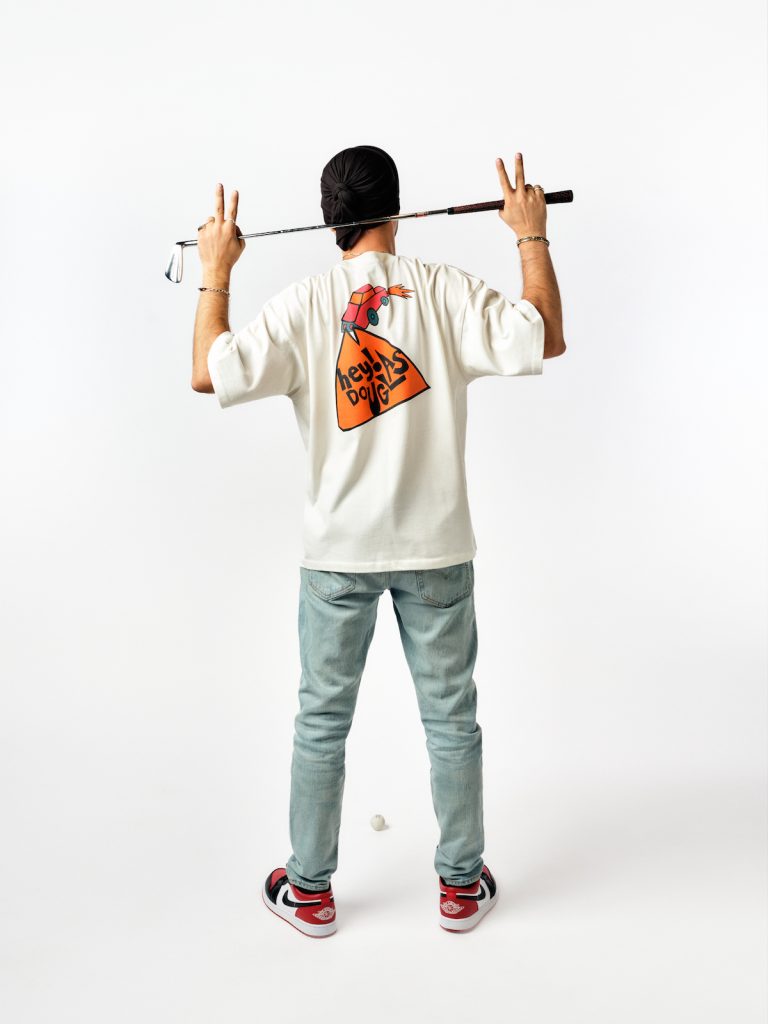 Hey! Douglas, İstanbul Çorbası koleksiyonu fotoğraf çekiminde. Üstünde beyaz renkli bir tişört var. Sırtı dönük. Elleri arasında bir golf sopası var.