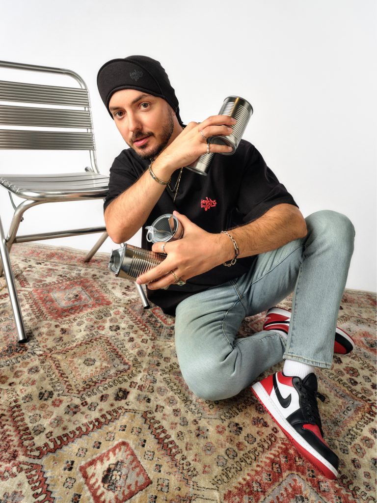 Hey! Douglas, İstanbul Çorbası koleksiyonu fotoğraf çekiminde. Üstünde siyah renkli bir tişört var. Yerde oturarak metal bir sandalyeye yaslanıyor. Elinde konserve kutusu açılmış, diğer elinde kağıyı tutuyor.