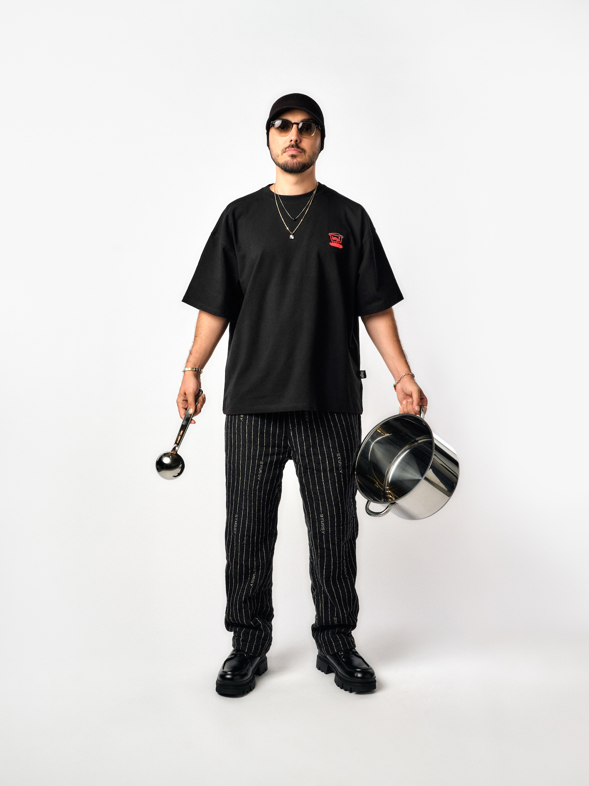 Hey! Douglas, İstanbul Çorbası koleksiyonu fotoğraf çekiminde. Üstünde siyah renkli bir tişört var. Koleksiyon adını betimlemek için bir elinde tencere, diğer elinde kepçe var.